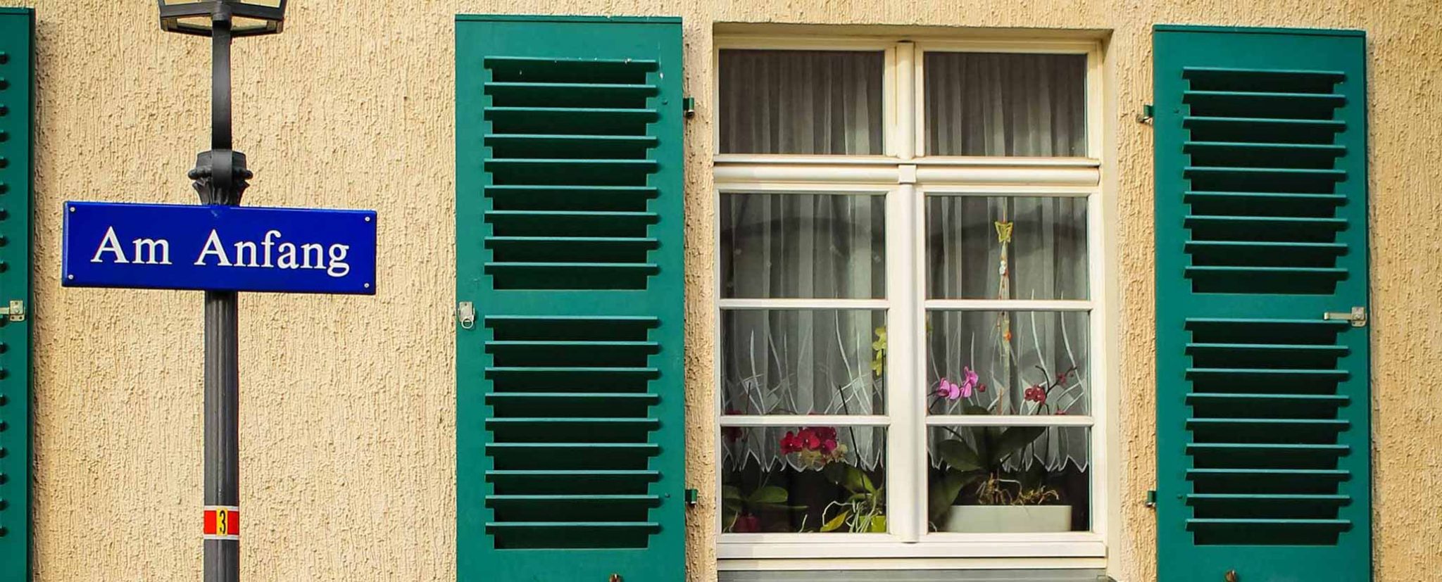 Eine Hausfassade mit grünen Fensterläden, davor ein Straßenschild mit der Aufschrift "Am Anfang"